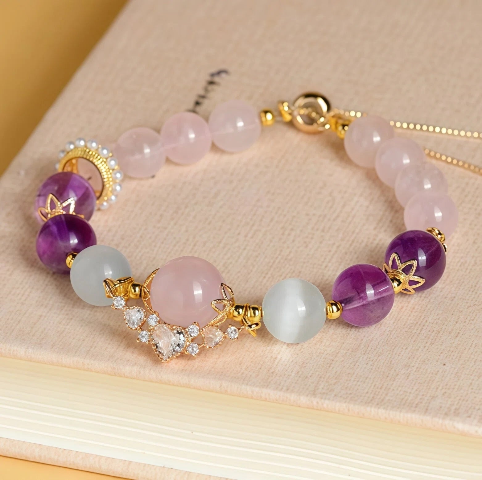 Styled flat lay of Cancer Zodiac Bracelet resting on a book. Featuring Rose Quartz, Cat's Eye, and Amethyst beads with a jeweled Crown charm for emotional healing - Kosmos Zen